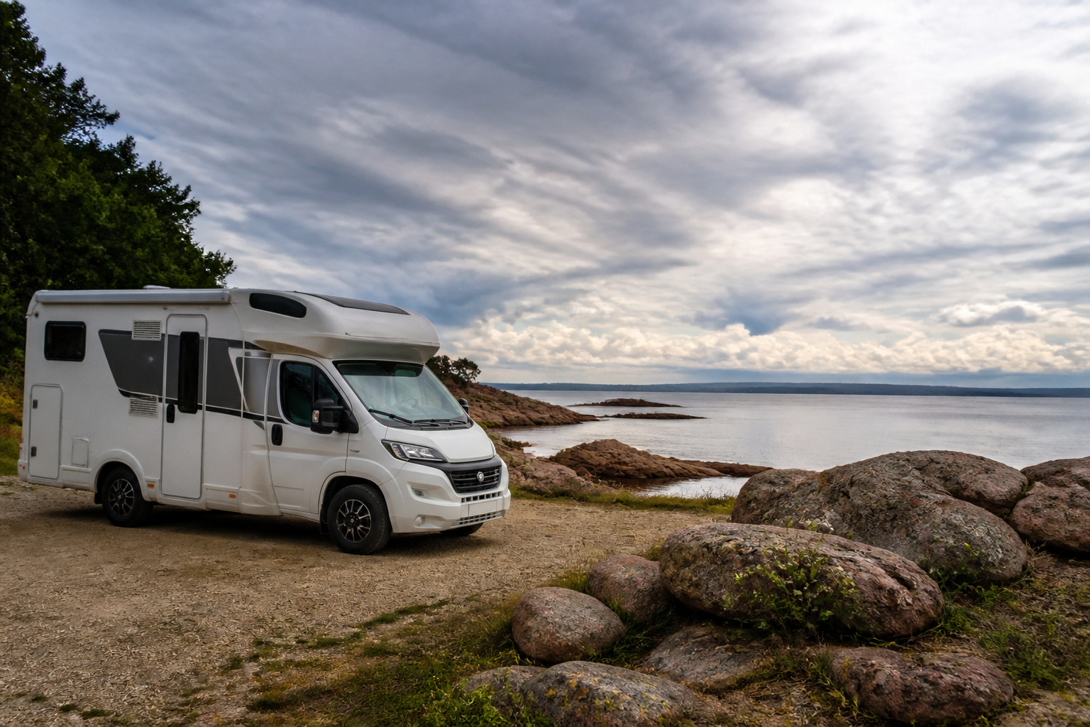 Un camping-car de taille moyenne garé sur une aire côtière en Bretagne, face à une baie découpée avec des rochers granitiques roses au premier plan et un ciel nuageux en arrière-plan