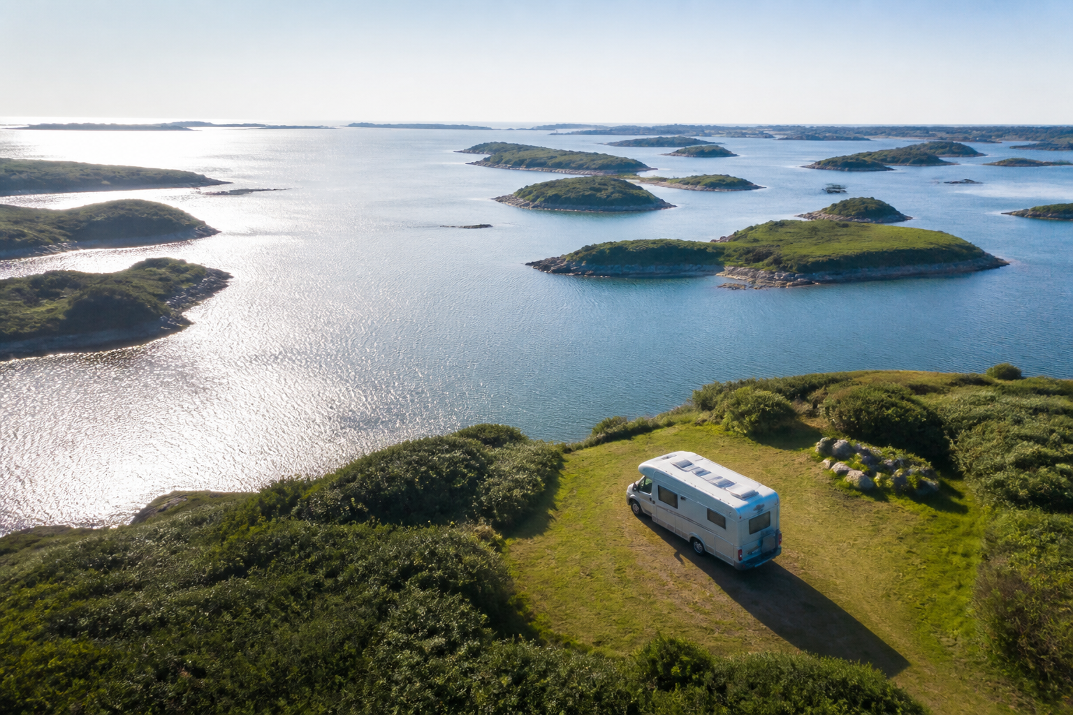 Vue aérienne d'un camping-car garé sur une aire herbeuse en bord de golfe en Bretagne, entouré de petites îles verdoyantes et d'une eau calme aux reflets argentés sous un ciel dégagé